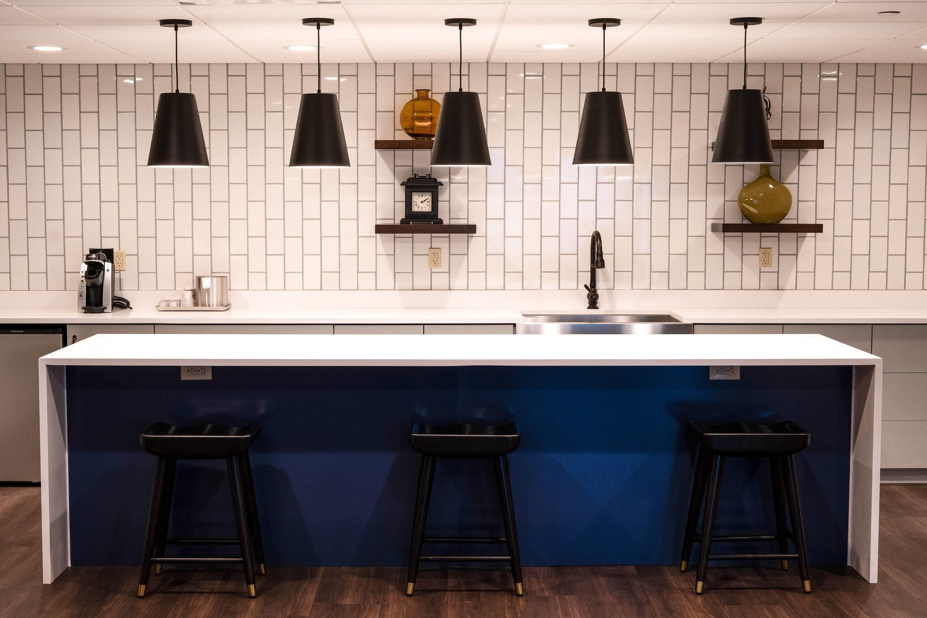 A modern kitchen with a white tiled backsplash, black pendant lights, a central island with stools, and a coffee machine.
