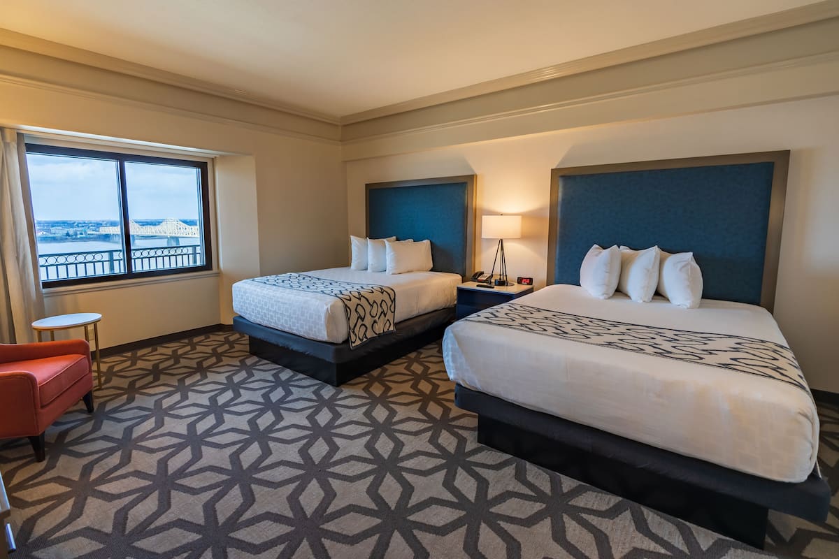 A hotel room with two beds, a red chair, a small table, and a view of a bridge.