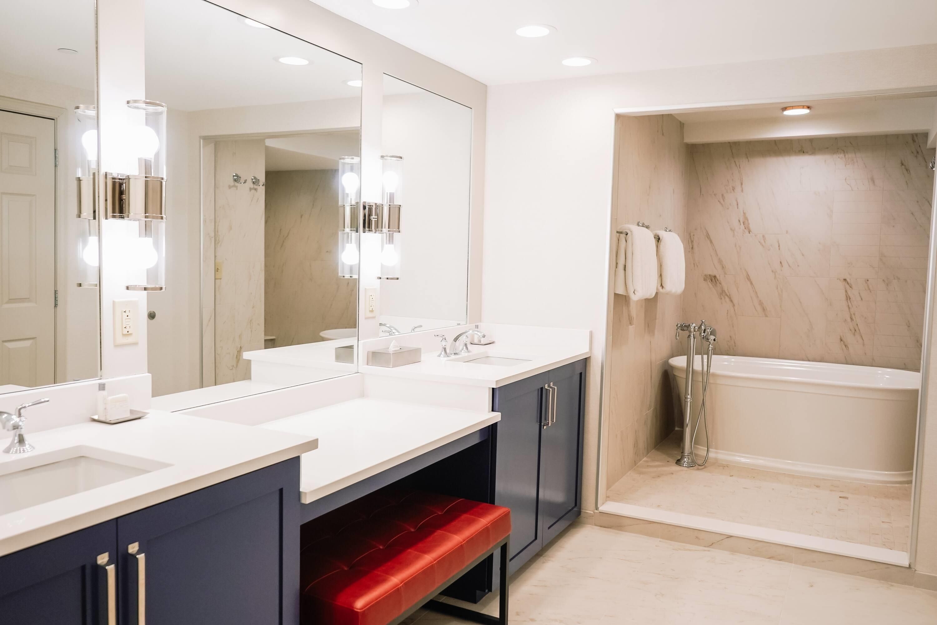A modern bathroom with a large mirror, two sinks, a red bench, a shower, and a bathtub.