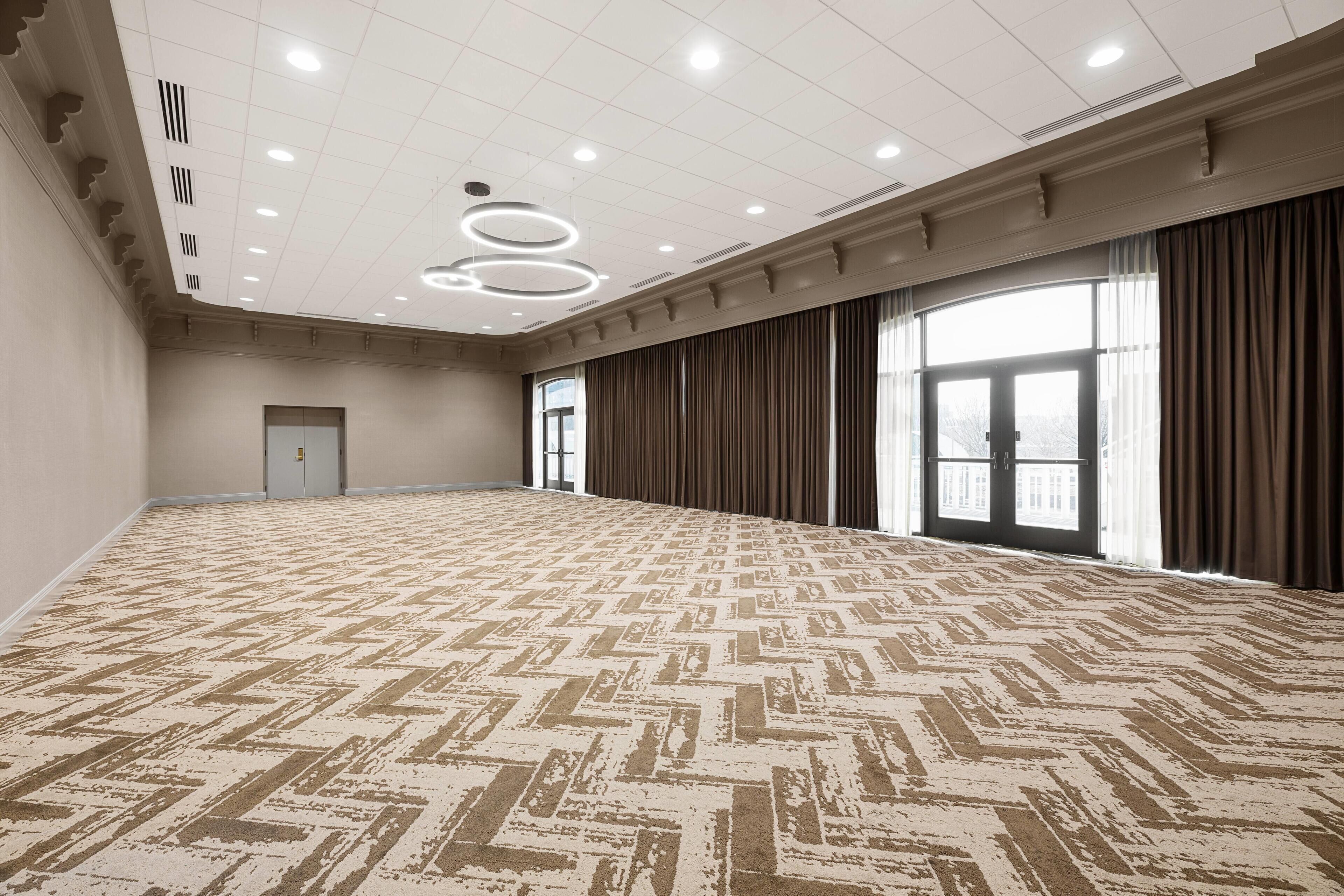 Un salón de conferencias amplio y vacío con alfombra estampada, techo alto con iluminación empotrada y un ventanal con cortinas.