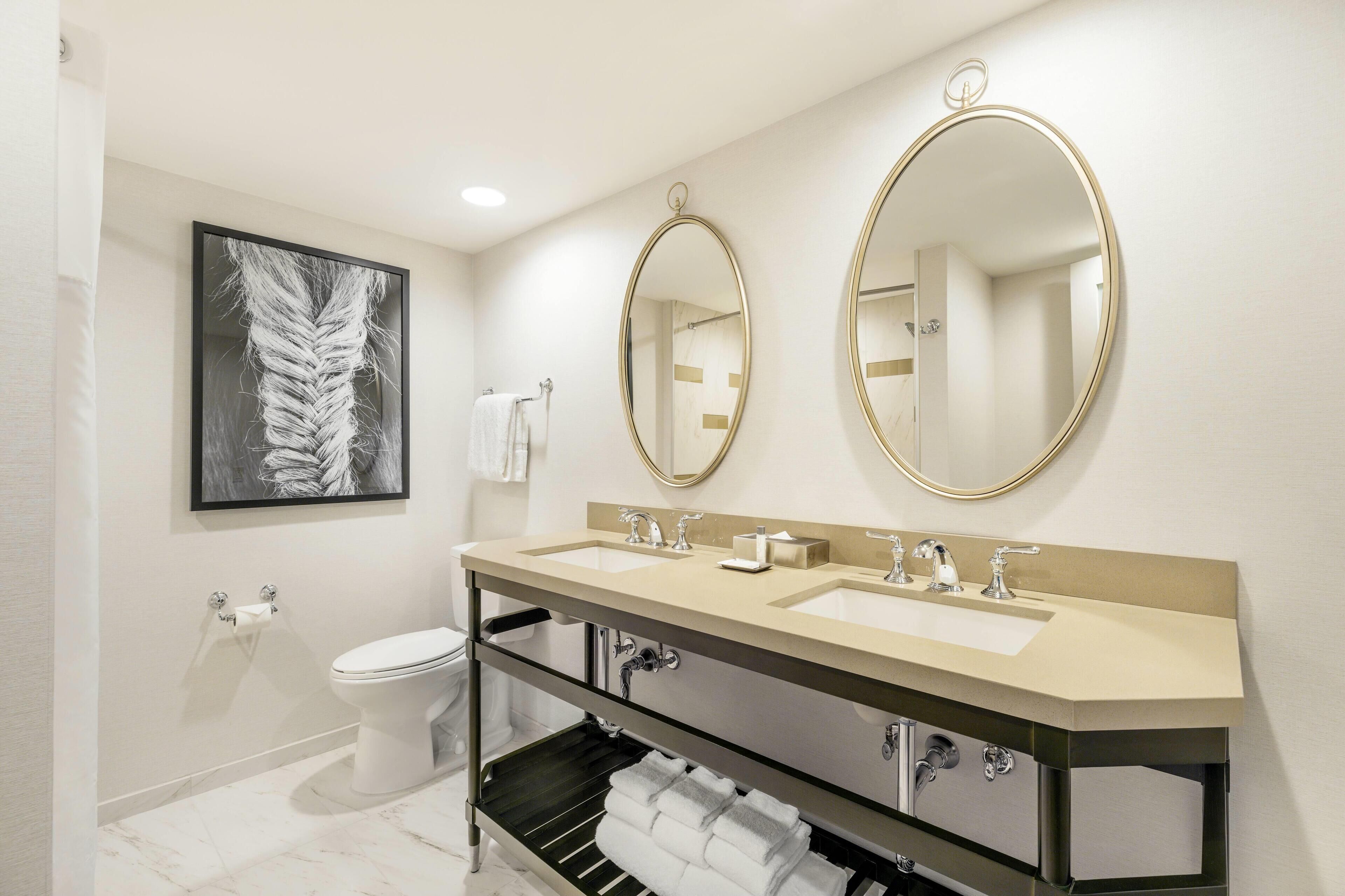 A modern bathroom with two sinks, a toilet, a towel rack, and a framed artwork on the wall.