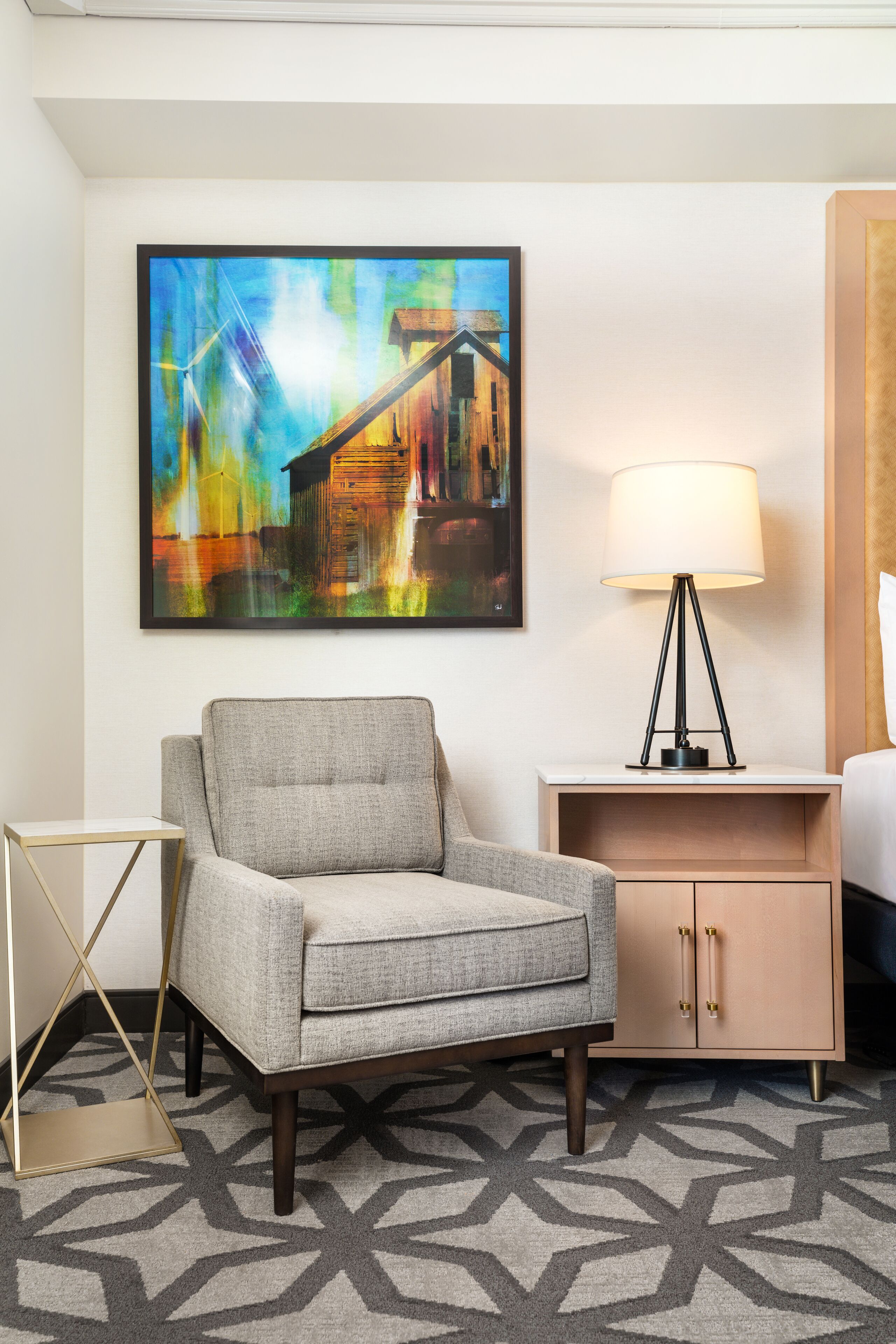 Una habitación de hotel con un sillón gris, una mesita de noche con lámpara, un mesita auxiliar de madera y un cuadro abstracto grande en la pared.