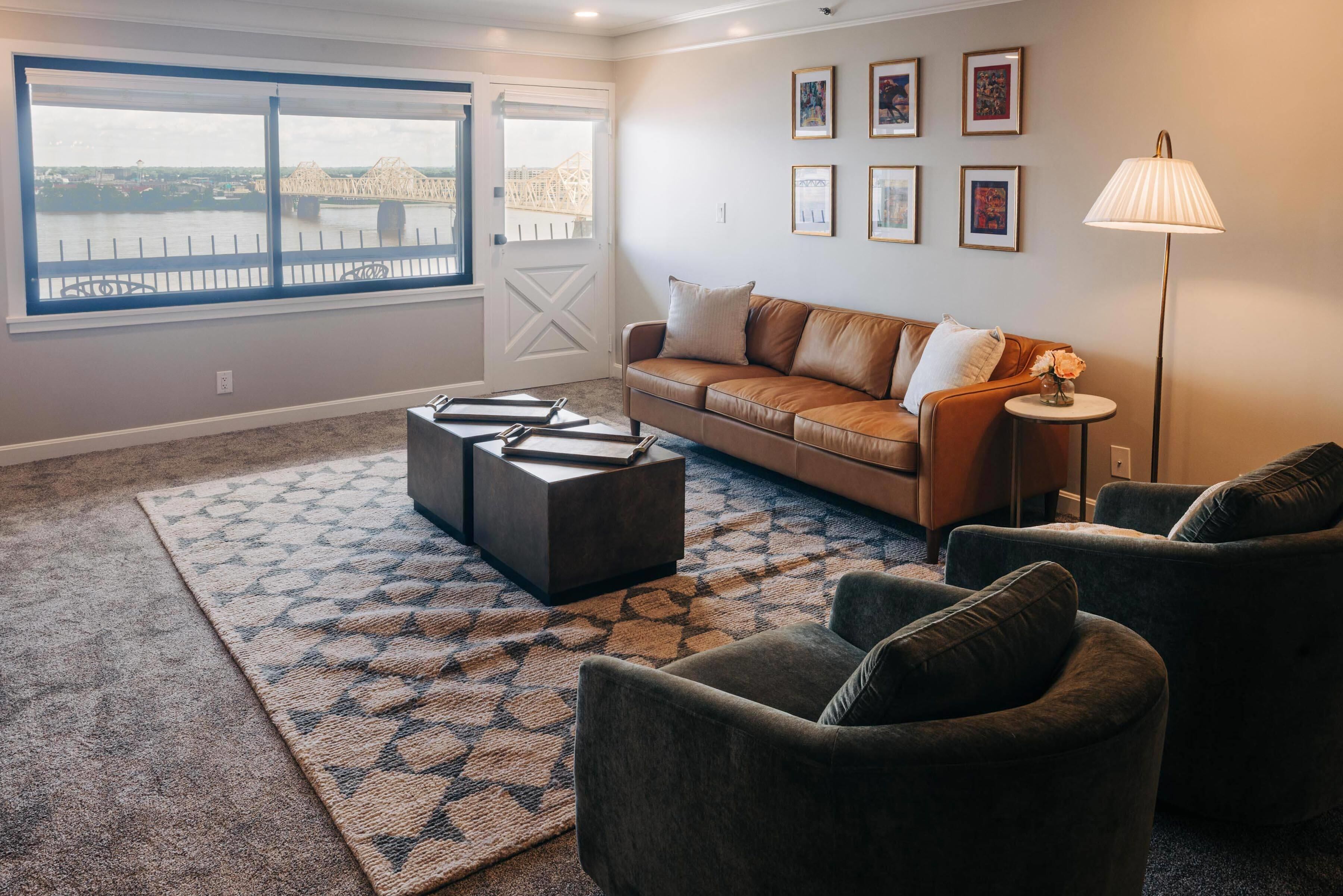 A living room with a large window, a brown leather sofa, a patterned rug, and a floor lamp.