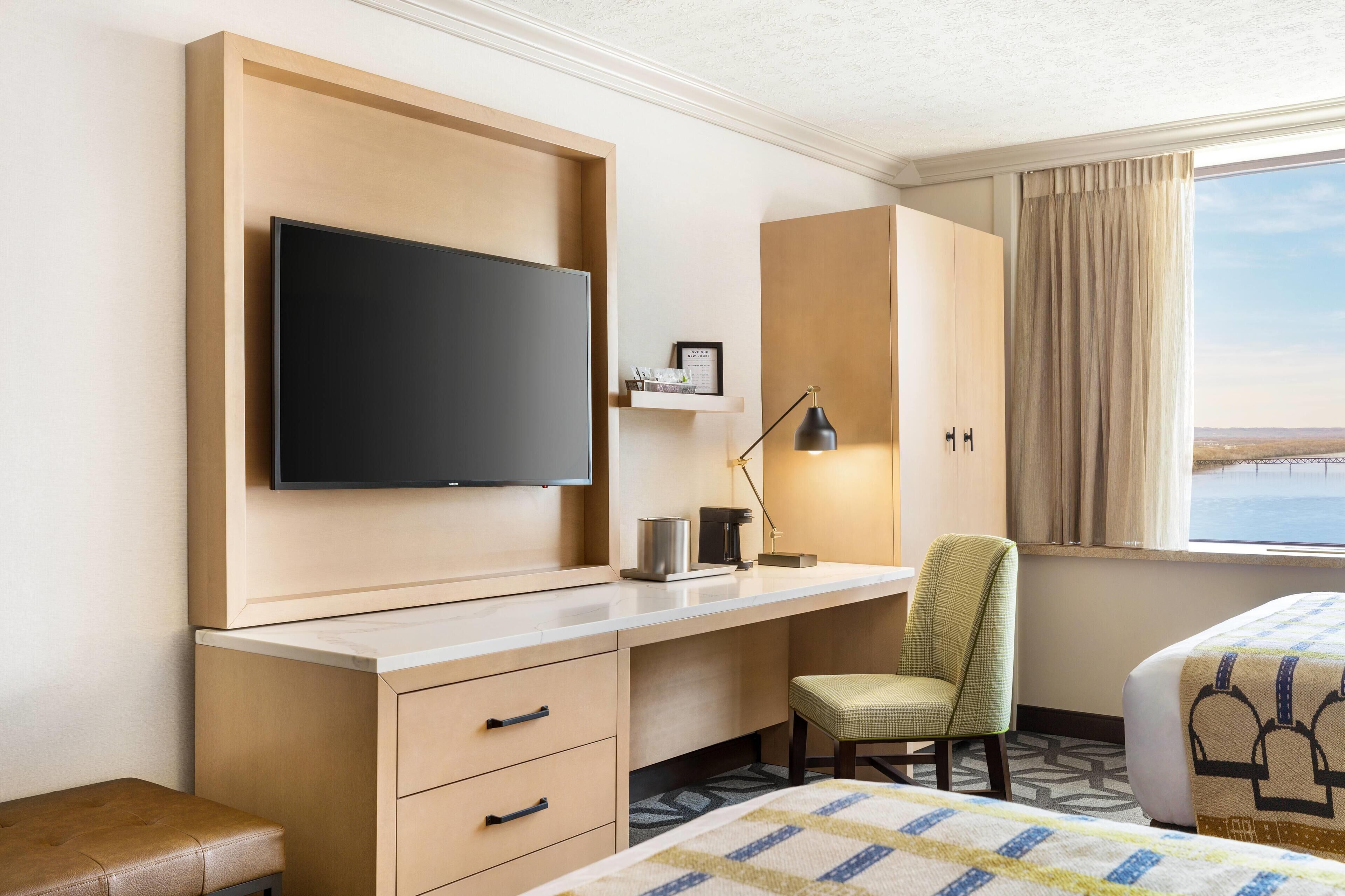 Habitación de hotel con un televisor de pantalla plana grande montado en la pared, un escritorio con lámpara, una silla, una cama con cubrecama estampado y vista al mar por la ventana.