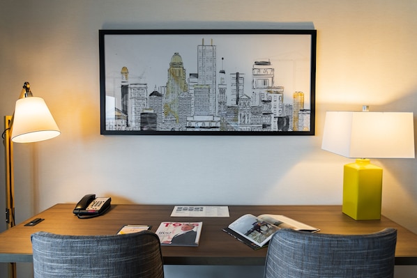Habitación de hotel con una obra de arte urbana en la pared, un escritorio con teléfono, una revista y una lámpara amarilla.