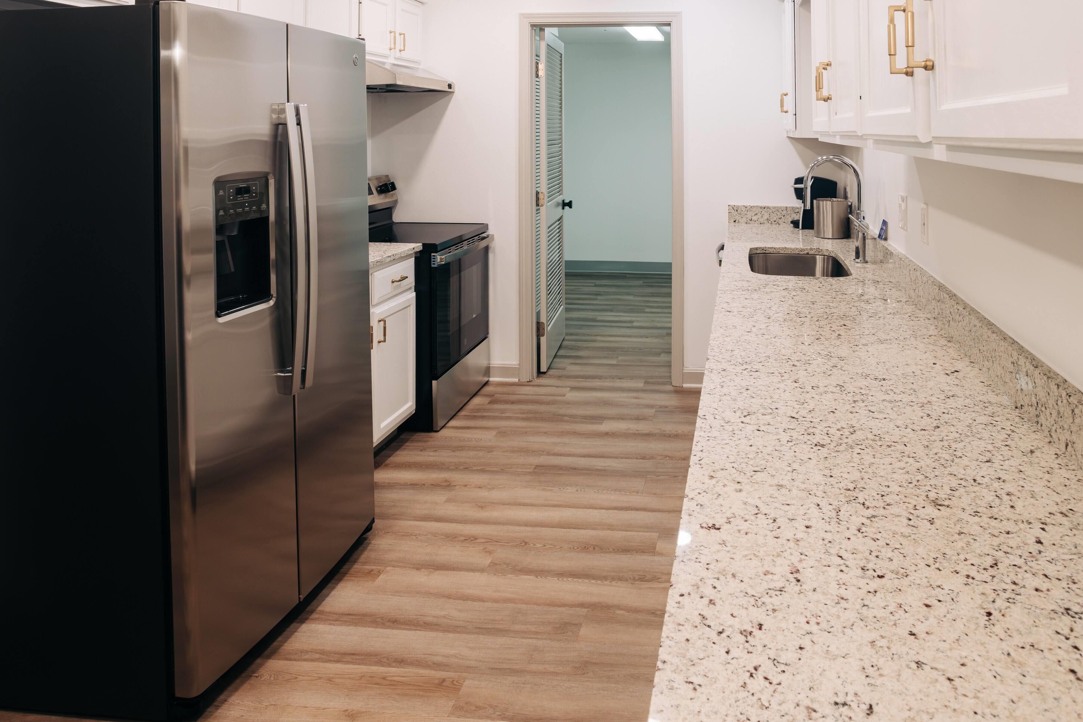 A kitchen with a stainless steel refrigerator, a built-in oven, a granite countertop, and a wooden floor.