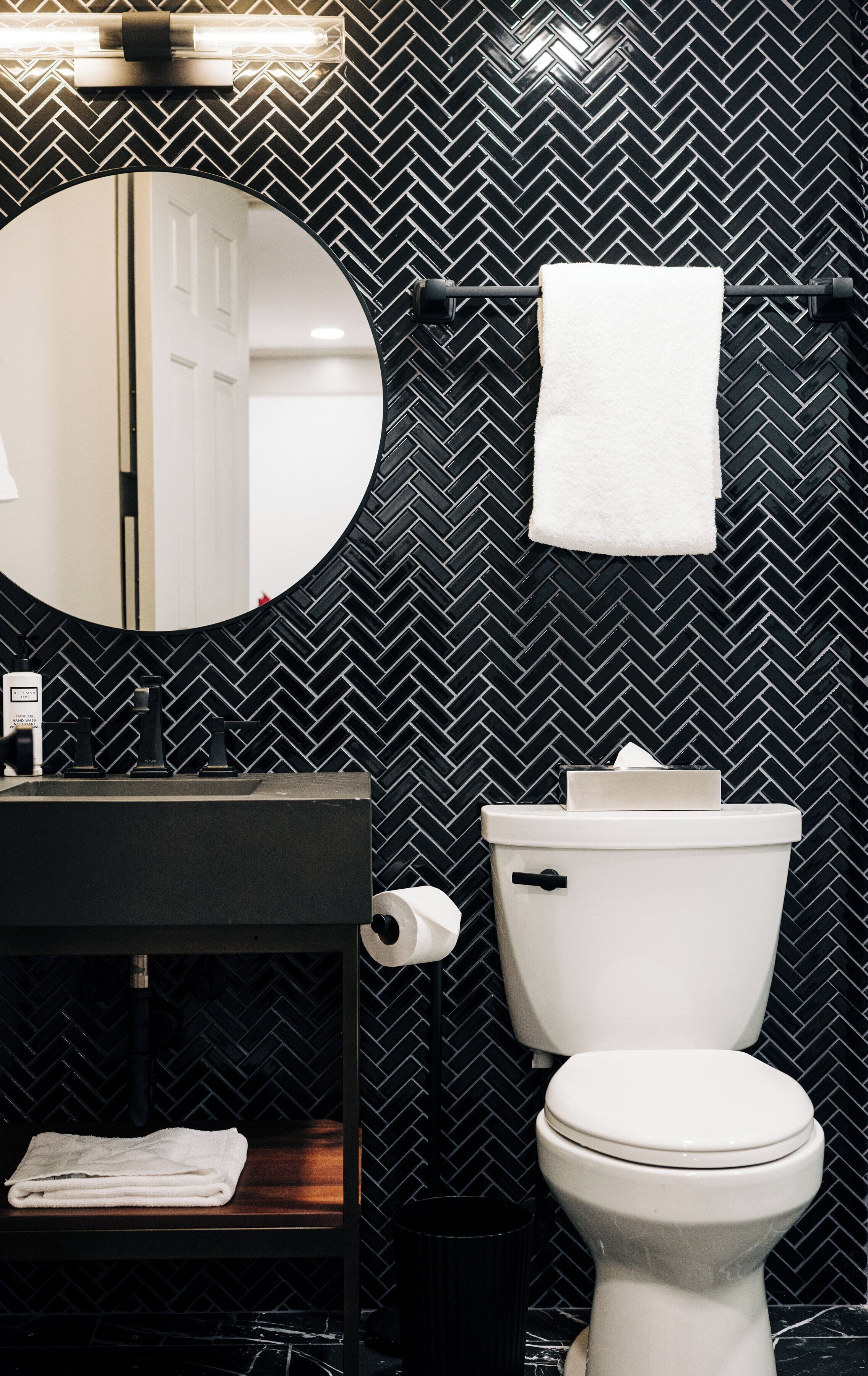 A bathroom with a black and white herringbone tile backsplash, a round mirror, a toilet, and a sink with a black countertop.