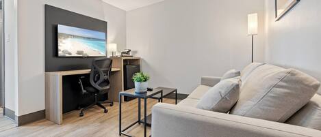 A modern hotel room with a sofa, a desk with a chair, a flat-screen TV, and a small table with a plant.