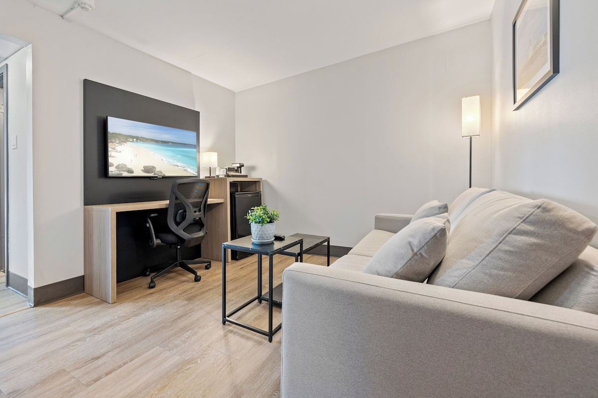 A modern hotel room with a wooden floor, a grey sofa, a desk with a computer, a TV mounted on the wall, and a bedside lamp.