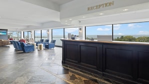 A reception area with a dark wood reception desk, large windows offering a view of the city, and a seating area with blue and red chairs. - The Jordan Ramada by Wyndham (Lincoln)