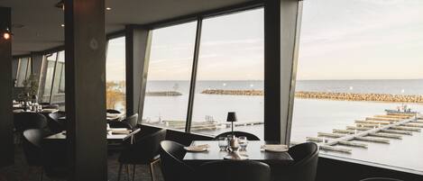 A restaurant with tables set for dining, large windows offering a view of the water and pier, and modern lighting fixtures.
