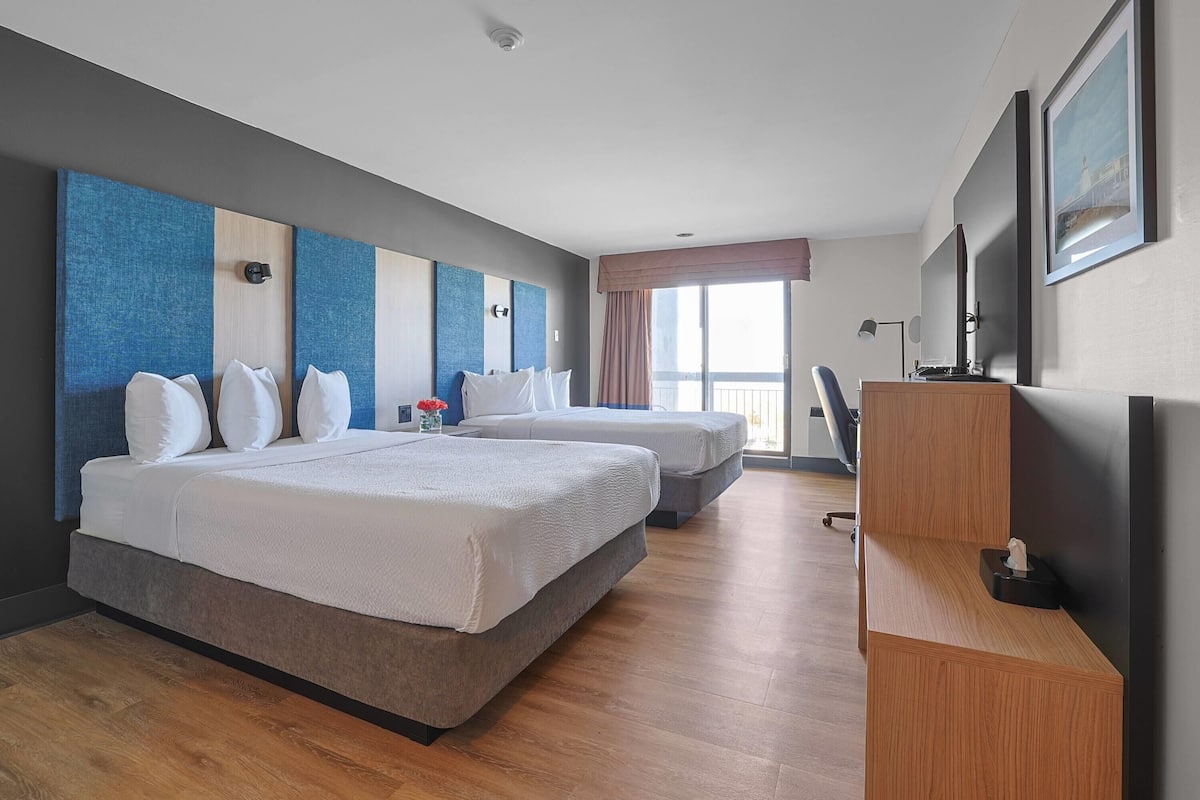 A hotel room with two beds, a desk, a chair, a TV, and a window with a view.