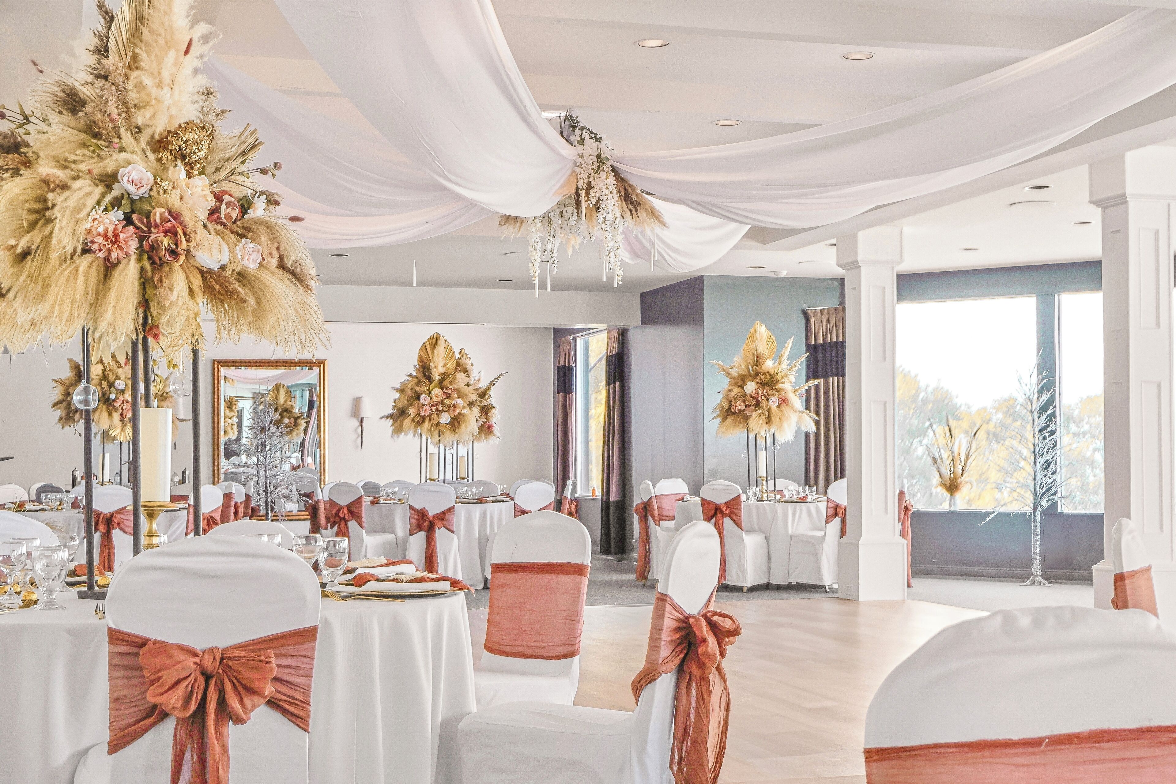 Uma sala de banquetes com mesas redondas, cadeiras brancas com fitas laranja e centros de mesa florais.