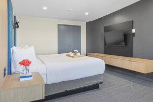 A modern hotel room with a large bed, a flat-screen TV mounted on the wall, a wooden nightstand with a book and a vase of flowers, and a bench along the wall.