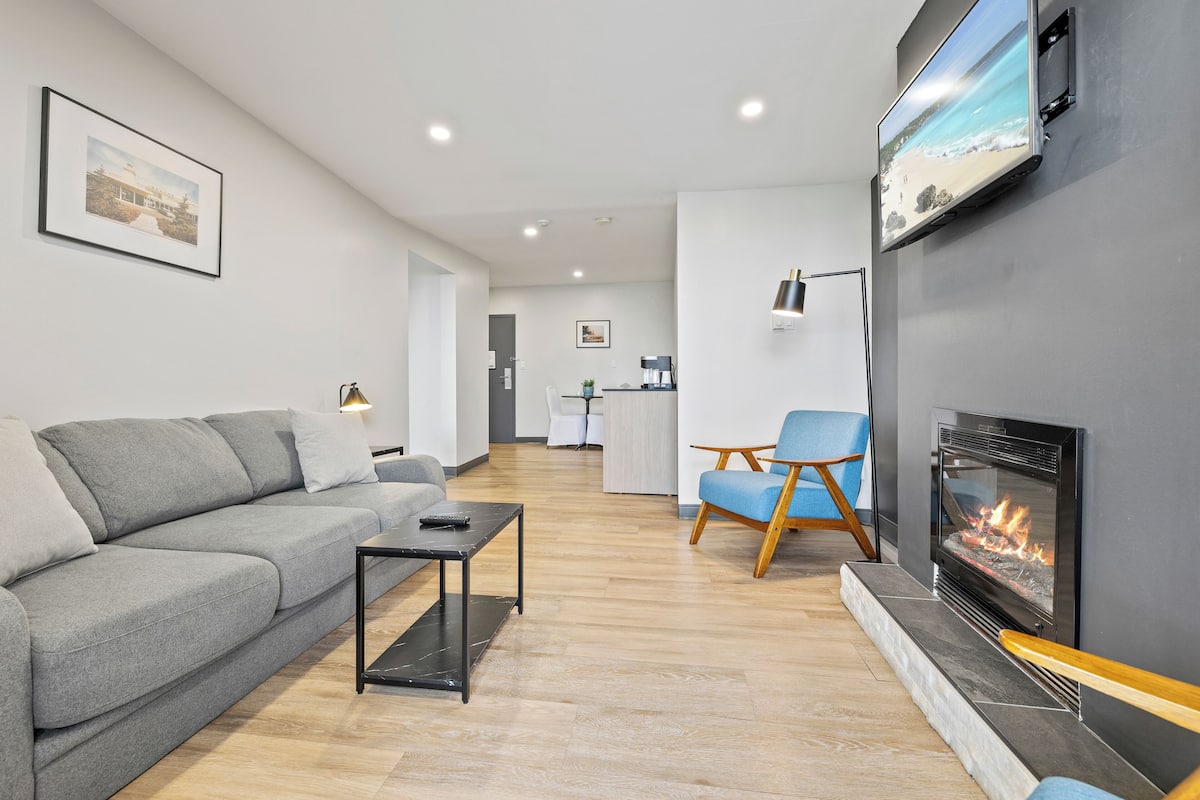 A modern living room with a grey sofa, a coffee table, a blue armchair, a fireplace, and a TV mounted on the wall.