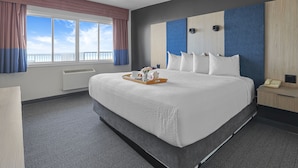 A hotel room with a large bed, a bedside table with a phone, a window with curtains, and a view of the sky. - The Jordan Ramada by Wyndham (Lincoln)