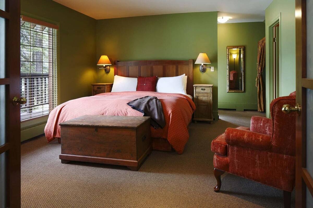 A bedroom with a large bed, two bedside lamps, a wooden chest, a red armchair, and a mirror on the wall.