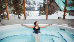 A person in a black swimsuit relaxing in a hot tub with a snowy mountain view in the background. - Emerald Lake Lodge (Field)