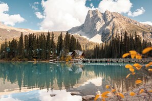 Una cabaña de montaña junto a un lago tranquilo con reflejos, rodeada de un bosque denso y una imponente cima montañosa.