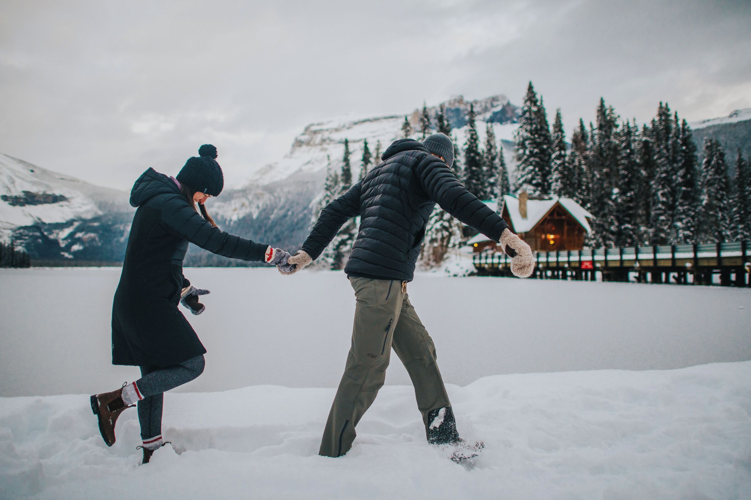 A couple holding hands in a snowy outdoor setting with a cabin in the background.