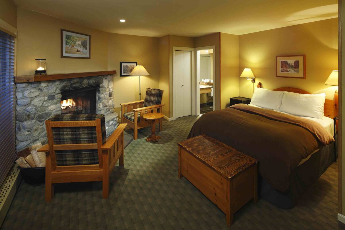 A hotel room with a stone fireplace, a bed with a brown bedspread, a wooden armchair, a small table, and a bench.