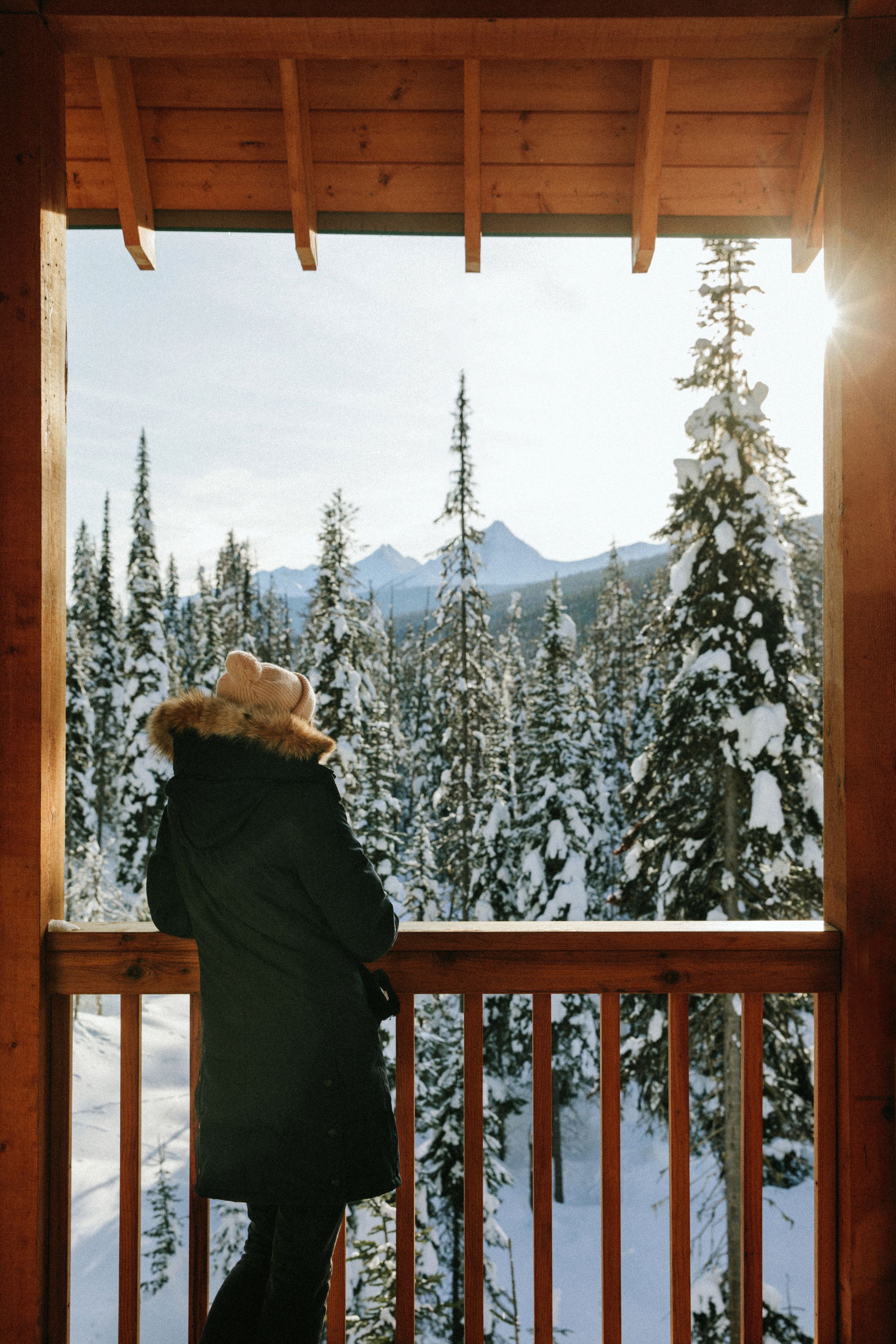 Eine Person steht auf einem Holz Balkon mit Blick auf einen verschneiten Wald und Berge im Hintergrund.
