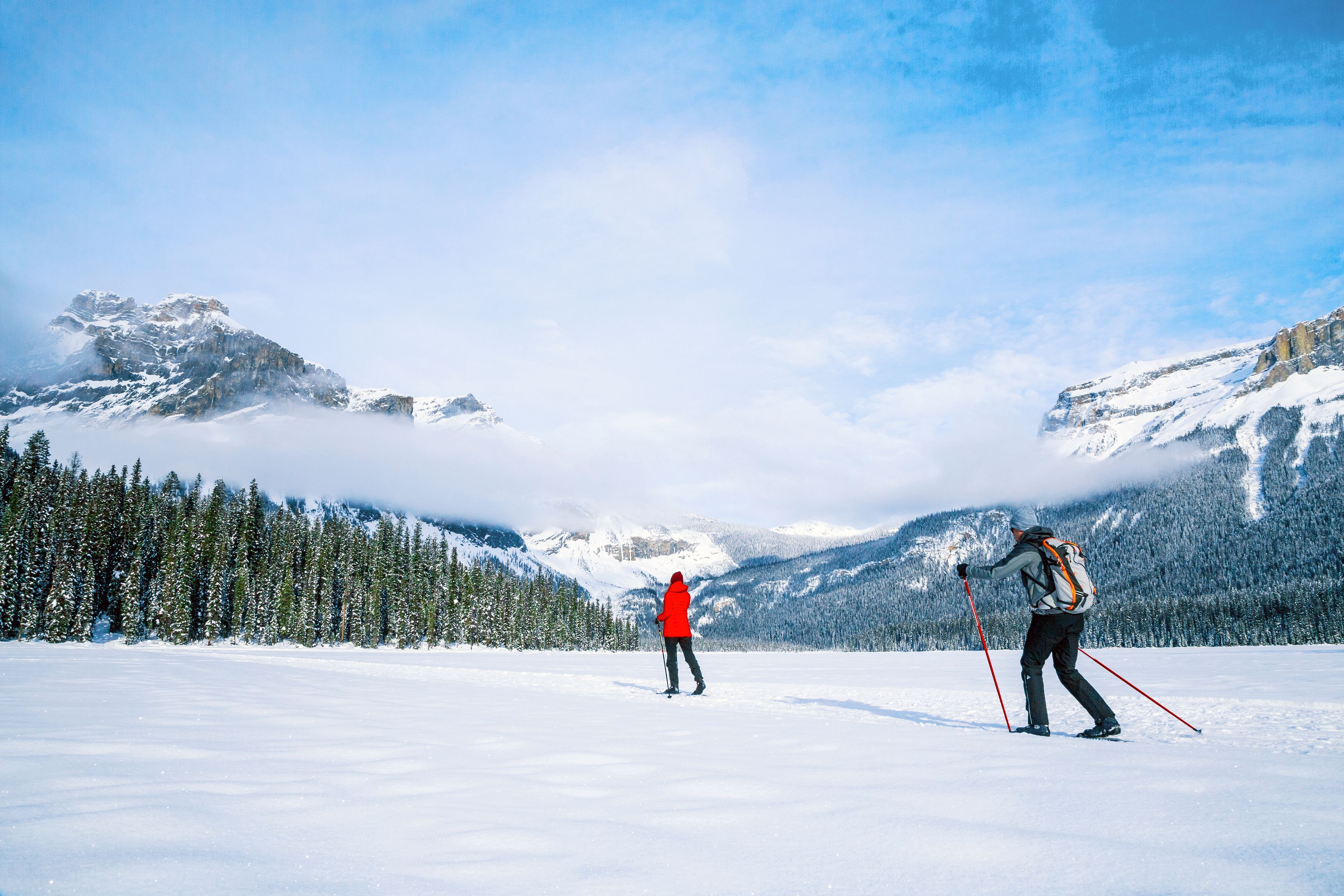 Schneelandschaft mit zwei Skiläufern, einer in einer roten Jacke und der andere in einer weißen Jacke, die auf einer ebenen, schneebedeckten Fläche entlanggehen, mit Bäumen und Bergen im Hintergrund.