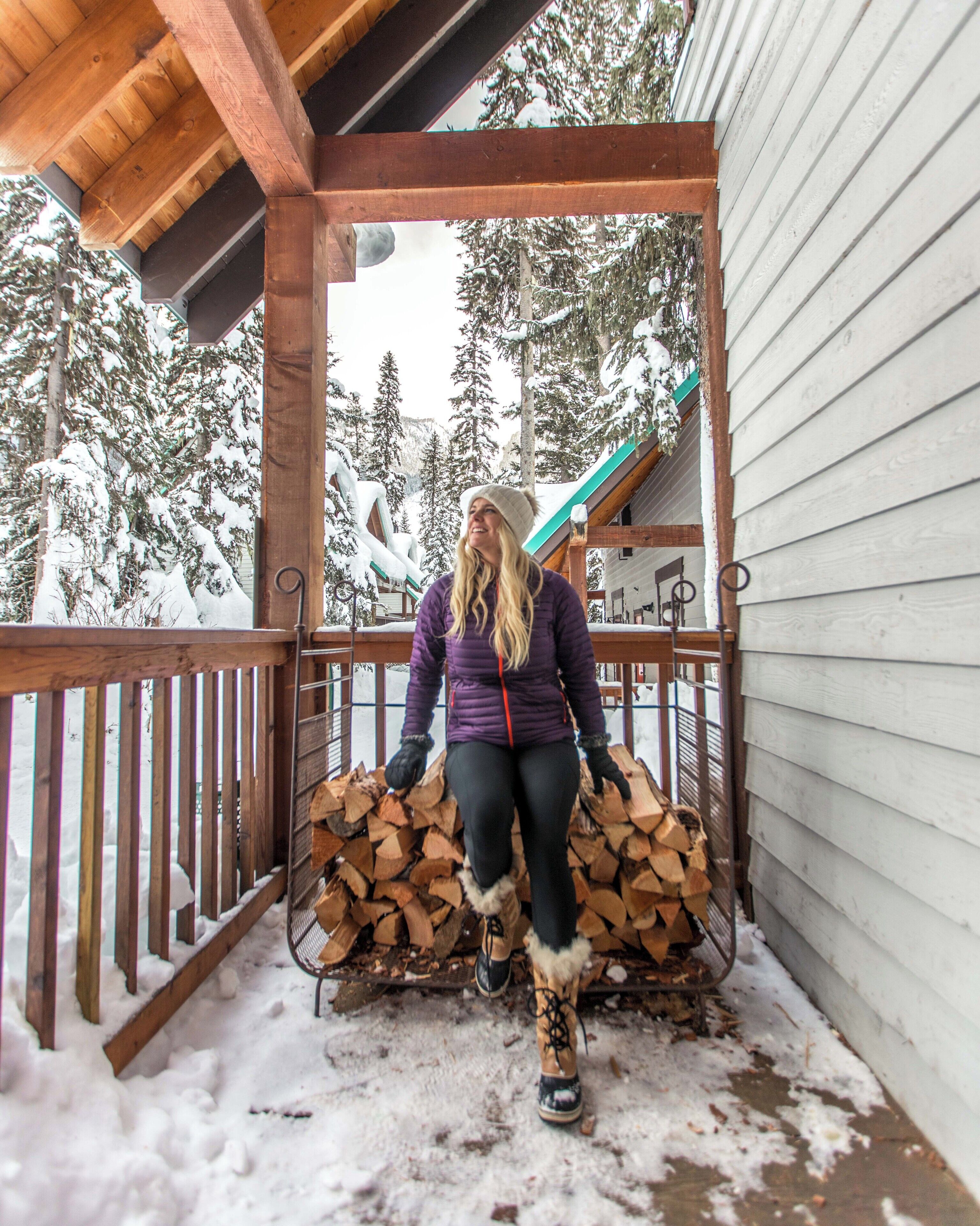 Una terraza de madera con baranda metálica, una pila de leña y árboles nevados al fondo.