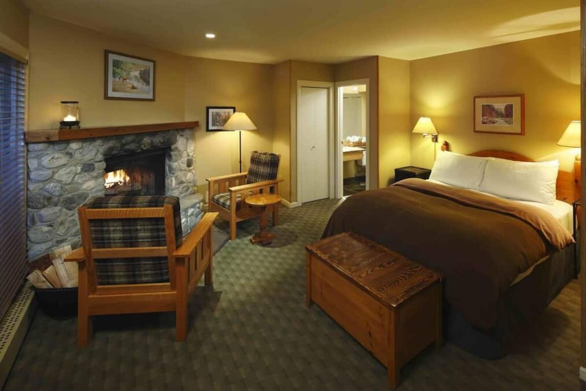 A hotel room with a stone fireplace, a bed with a brown blanket, a wooden chair, a small table, and a nightstand with a lamp.