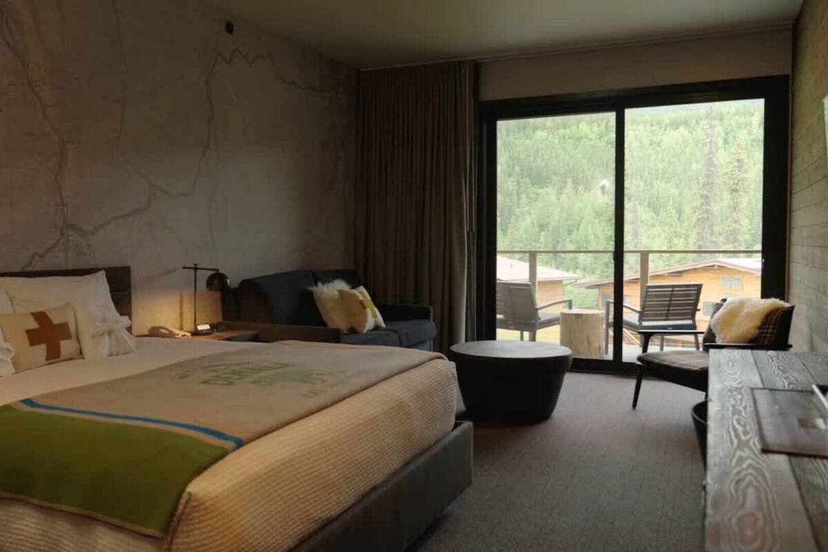 A hotel room with a large bed, a sofa, a small table, and a balcony with outdoor seating.