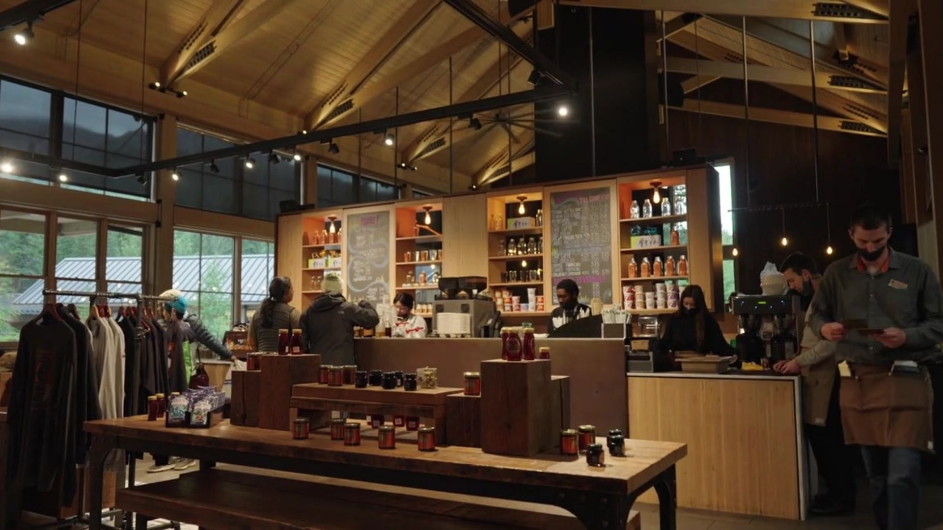 A café with wooden tables, shelves stocked with beverages, and a view of the outdoors through large windows.