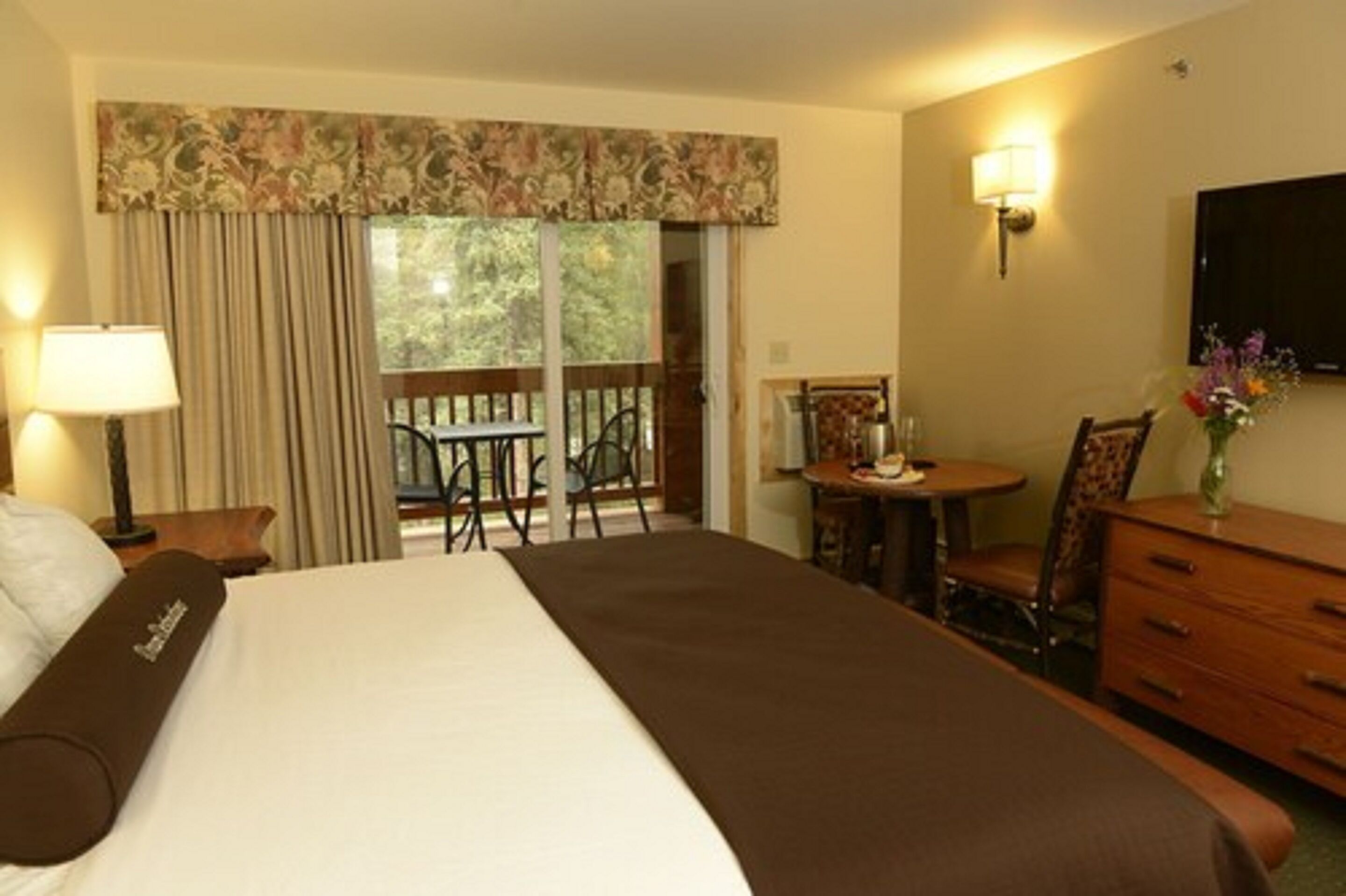 Una habitación de hotel con una cama grande, un escritorio con silla, una mesita con silla, un aparador, un televisor montado en la pared y una ventana con cortinas.