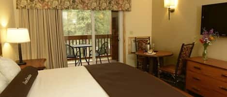 Una habitación de hotel con una cama grande, un escritorio con silla, una mesita con silla, un aparador, un televisor montado en la pared y una ventana con cortinas.