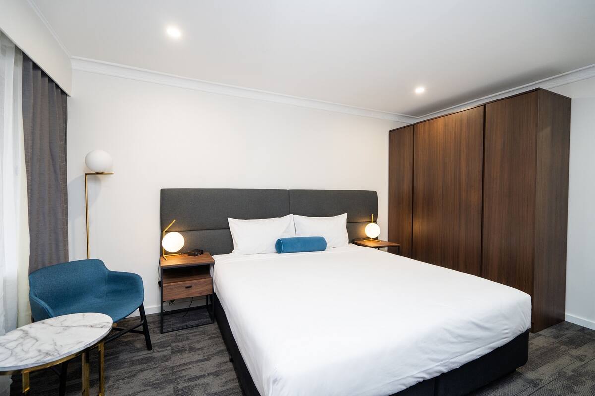 A modern hotel room with a large bed, a desk with a chair, a wardrobe, and a bedside table with lamps.