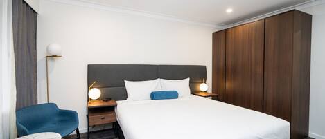 A modern hotel room with a large bed, a desk with a chair, a wardrobe, and a bedside table with lamps.