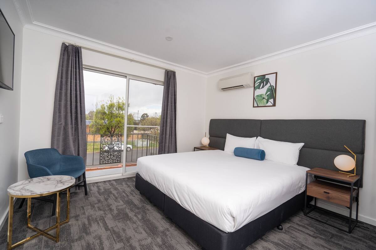 A hotel room with a large bed, a small table, a chair, and a view of the outdoors.