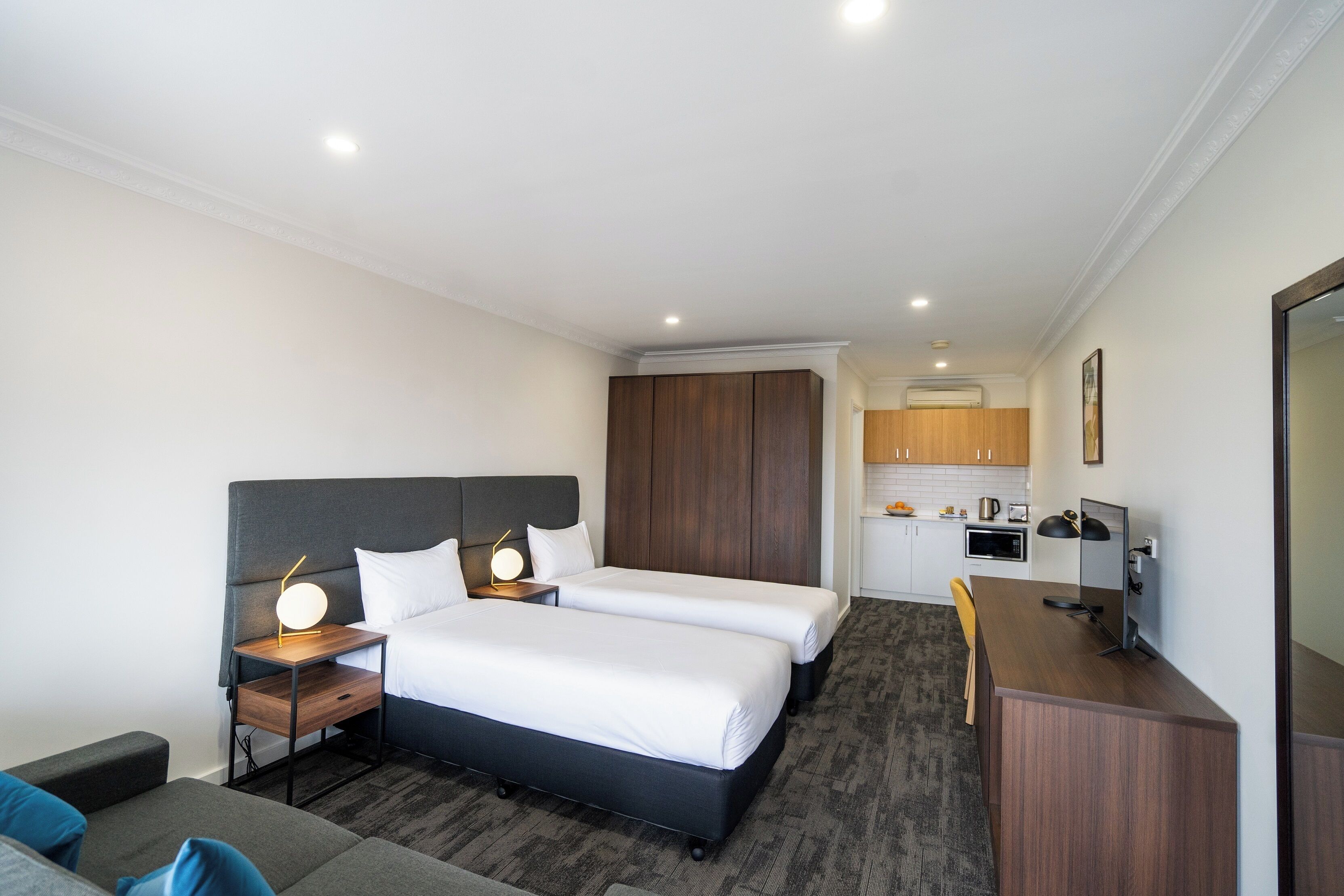 Habitación de hotel con dos camas, un escritorio, una silla, un pequeño sofá y una kitchenette al fondo.