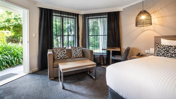 A hotel room with a bed, a sofa, a small table, and a chair. There is a window with curtains, a door, and a view of greenery outside. - Nightelier Doncaster (Doncaster)