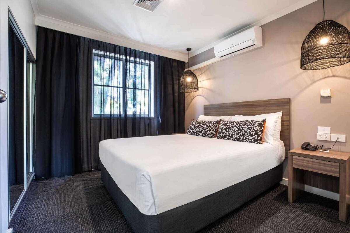 Ein Hotelzimmer mit einem großen Bett, einem Nachttisch mit Telefon, einer Wandklimaanlage und einem Fenster mit Vorhängen.