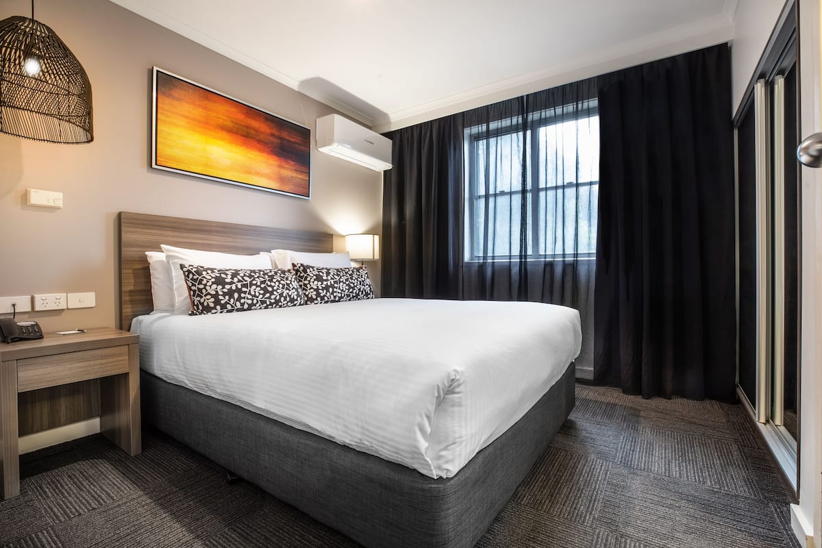 Ein Hotelzimmer mit einem großen Bett, einem Nachttisch mit Lampe, einer Wand-Klimaanlage und einem Fenster mit Vorhängen.