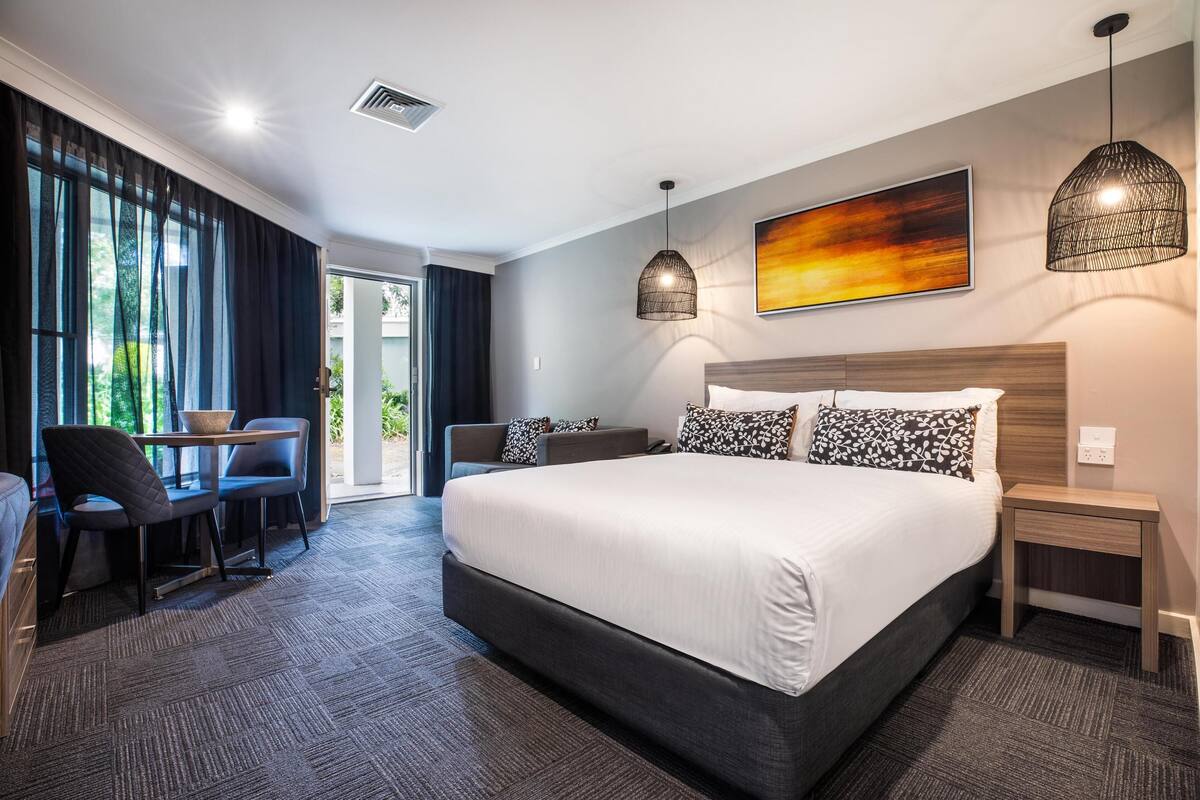 Ein modernes Hotelzimmer mit einem großen Bett, einem Schreibtisch mit zwei Stühlen, einem kleinen Tisch und Blick auf Grün hinter den Fenstern.