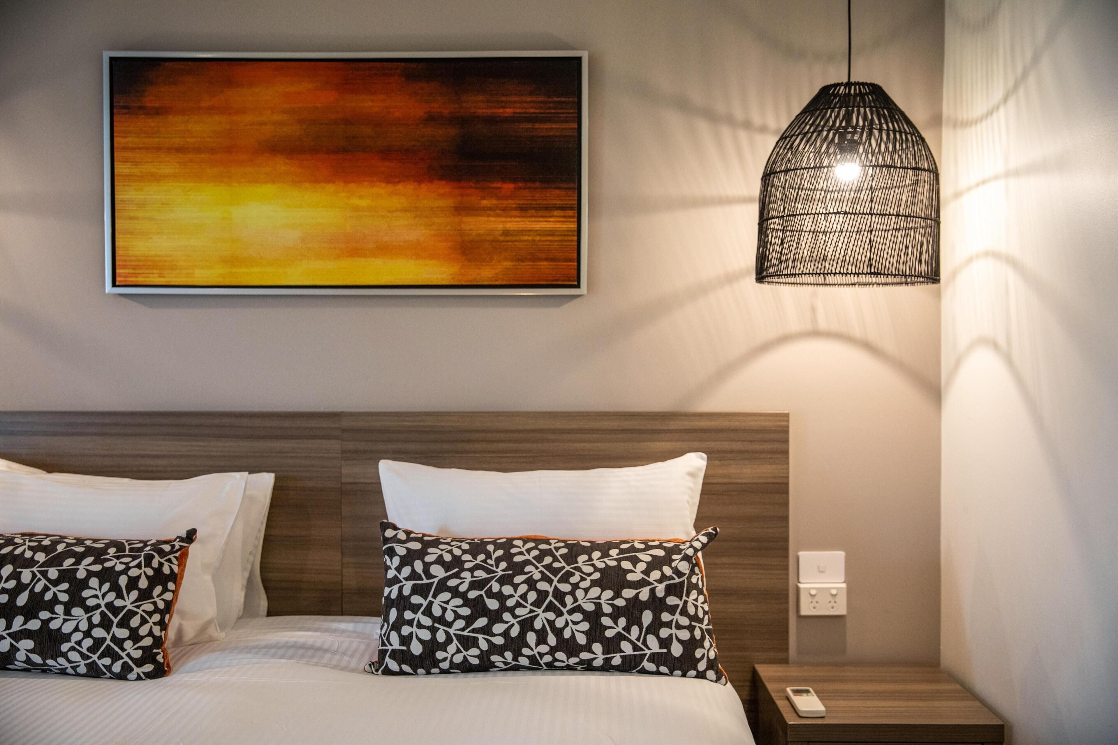 Een keurig opgemaakt bed met witte en gemêleerde kussens, een groot abstract schilderij aan de muur en een hanglamp.