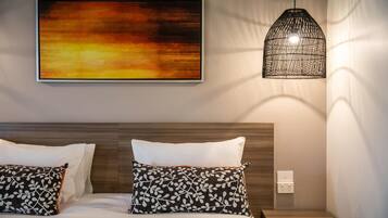 Een keurig opgemaakt bed met witte en gemêleerde kussens, een groot abstract schilderij aan de muur en een hanglamp.