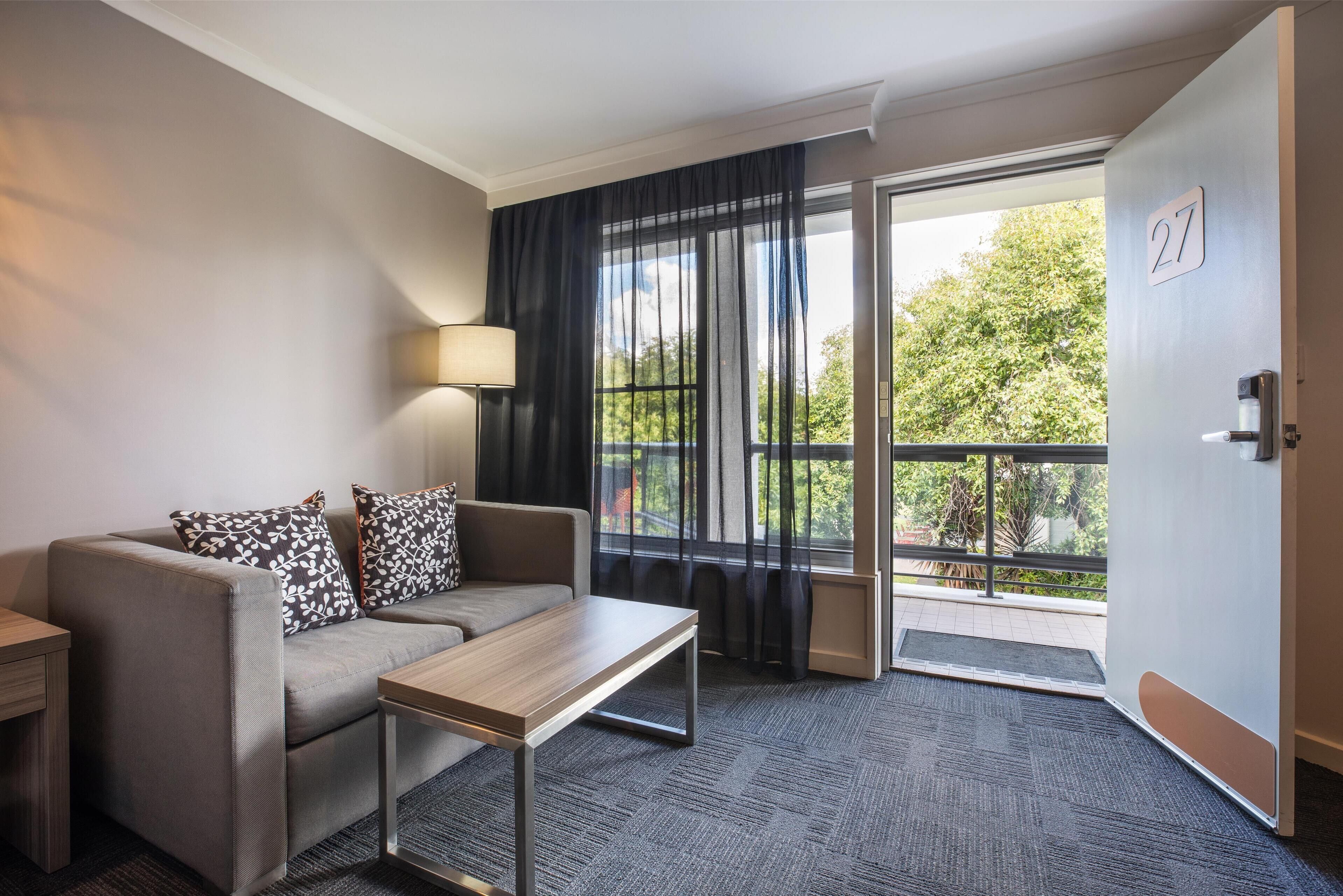 A hotel room with a sofa, a small table, a lamp, a sliding glass door to an outdoor balcony, and a door with the number 27.