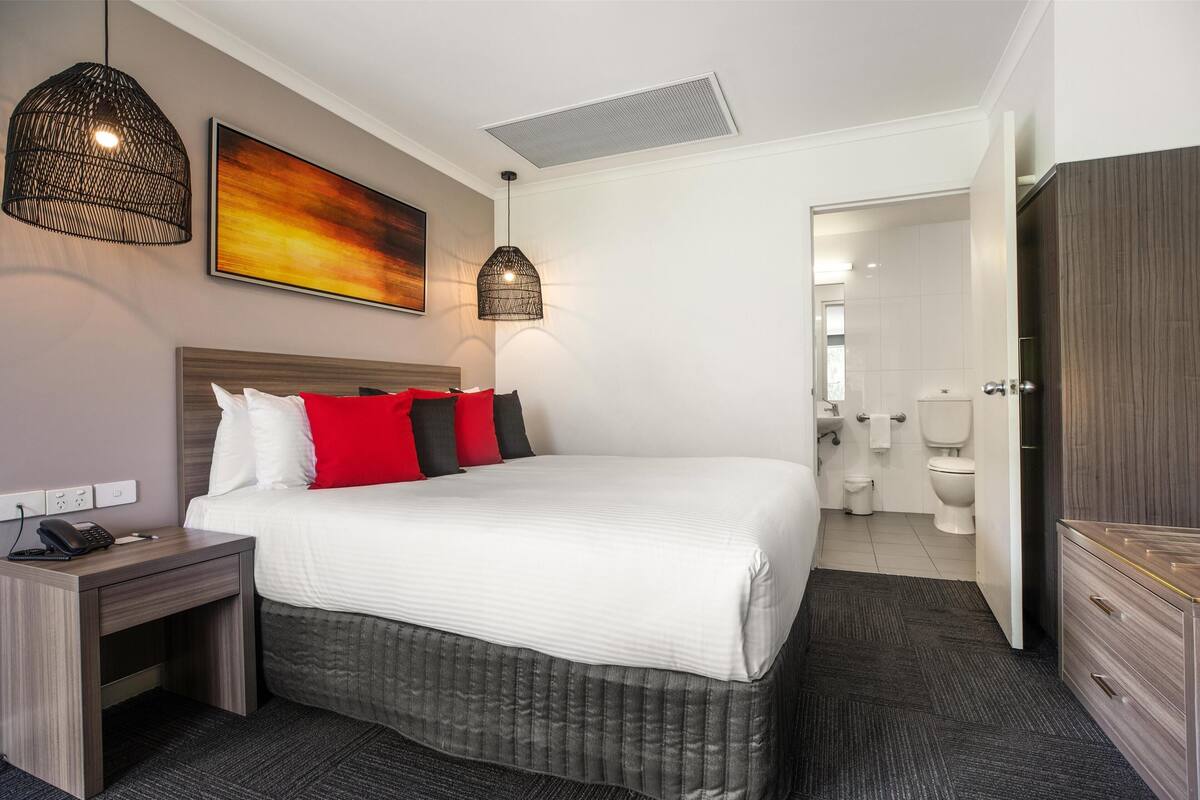 Ein Hotelzimmer mit einem großen Bett, einem Nachttisch mit Telefon, zwei Pendelleuchten, einem Bild an der Wand und Blick ins Badezimmer.