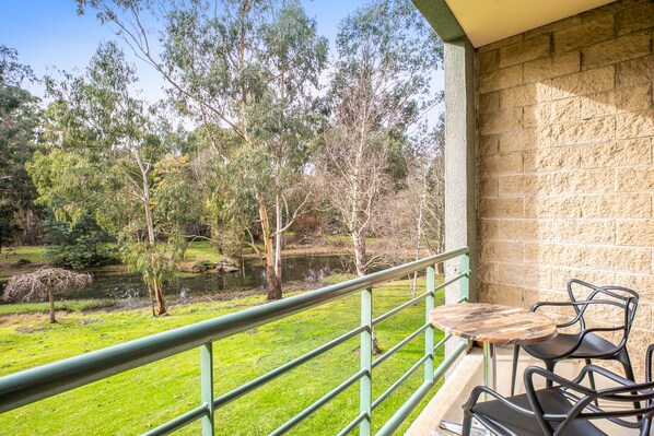 A balcony with a view of a pond and trees, a stone wall, and two metal chairs.