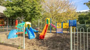 Un parque infantil con toboganes y estructuras para escalar, rodeado por una valla y árboles.