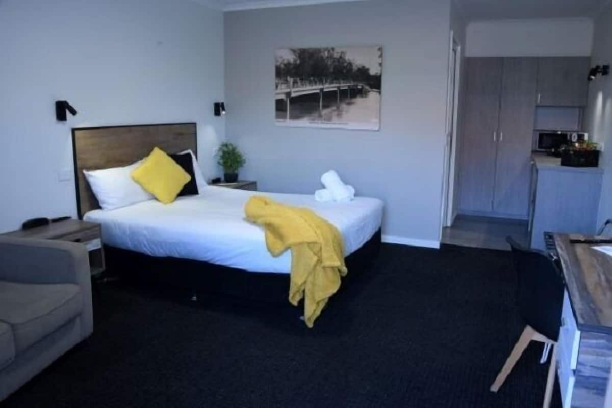 A hotel room with a bed, a bedside table, a chair, a desk, and a kitchenette.