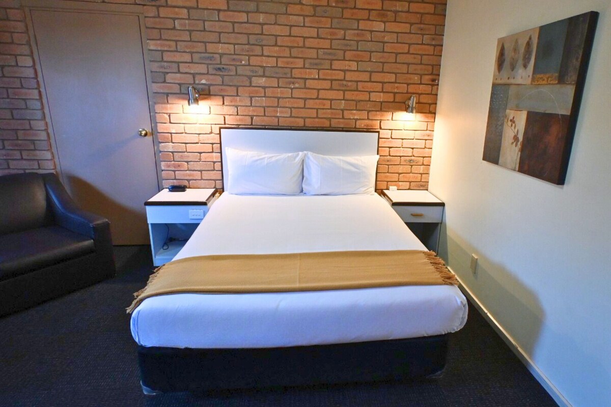 A hotel room with a brick wall, a bed with white bedding, two bedside tables, a nightstand, a chair, and a painting on the wall.