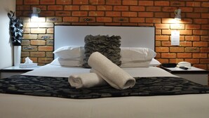 A neatly made bed with white linens, a rolled towel, and a decorative pillow. There is a nightstand with a lamp and a phone. The room has a brick wall and a decorative wall piece. - Peppermill Inn (Kialla)
